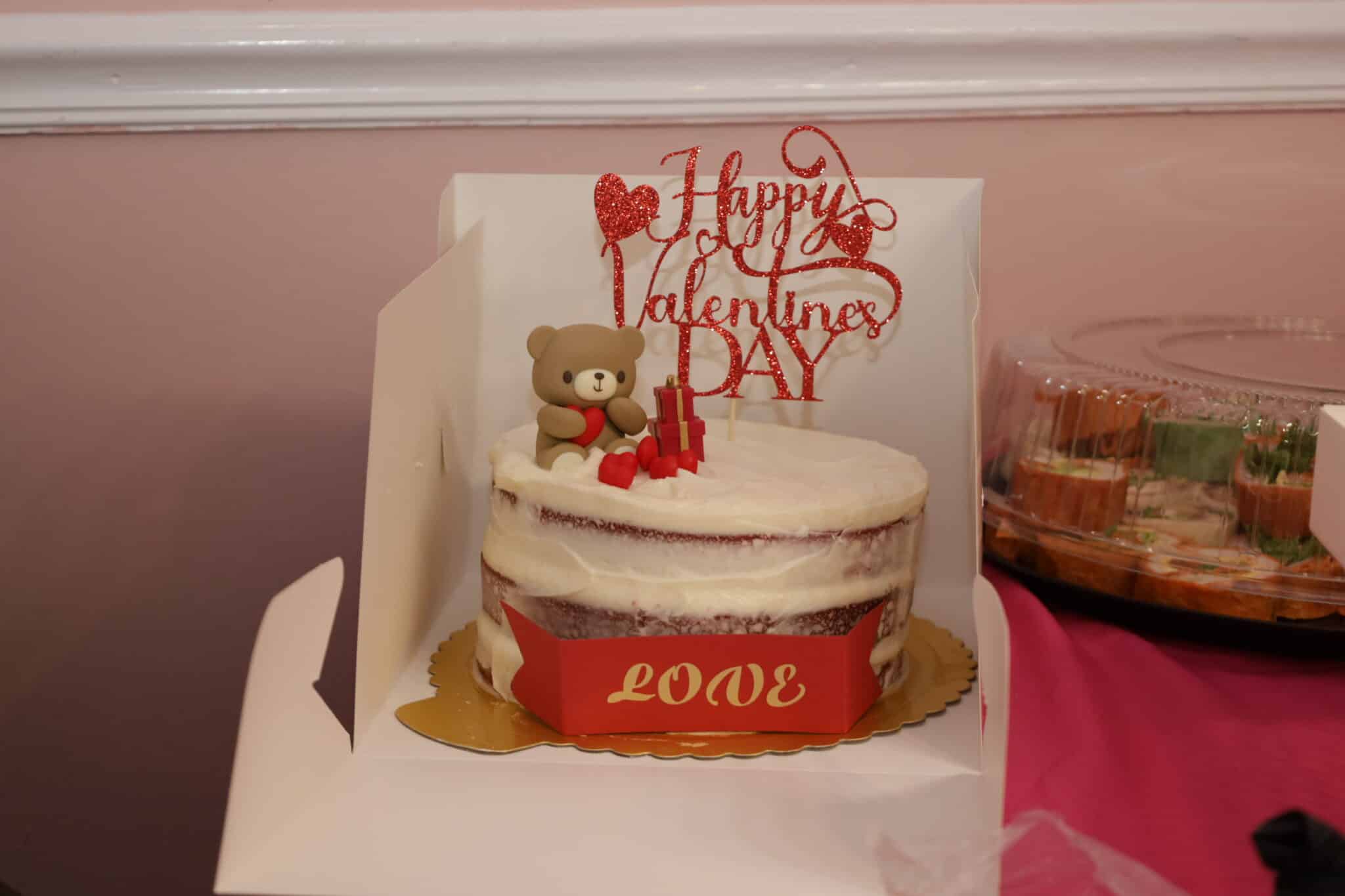 A white cake with a topper reading "Happy Valentine's Day" along with a brown teddy bear and a banner that reads "LOVE"