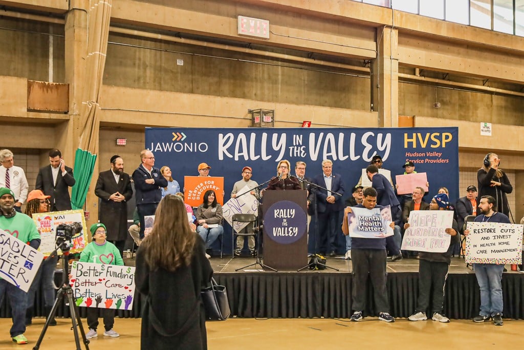 A group of people, including those supported by AHRC Nassau and staff members, stand at the podium with signs at a rally as a speaker gives a speech.